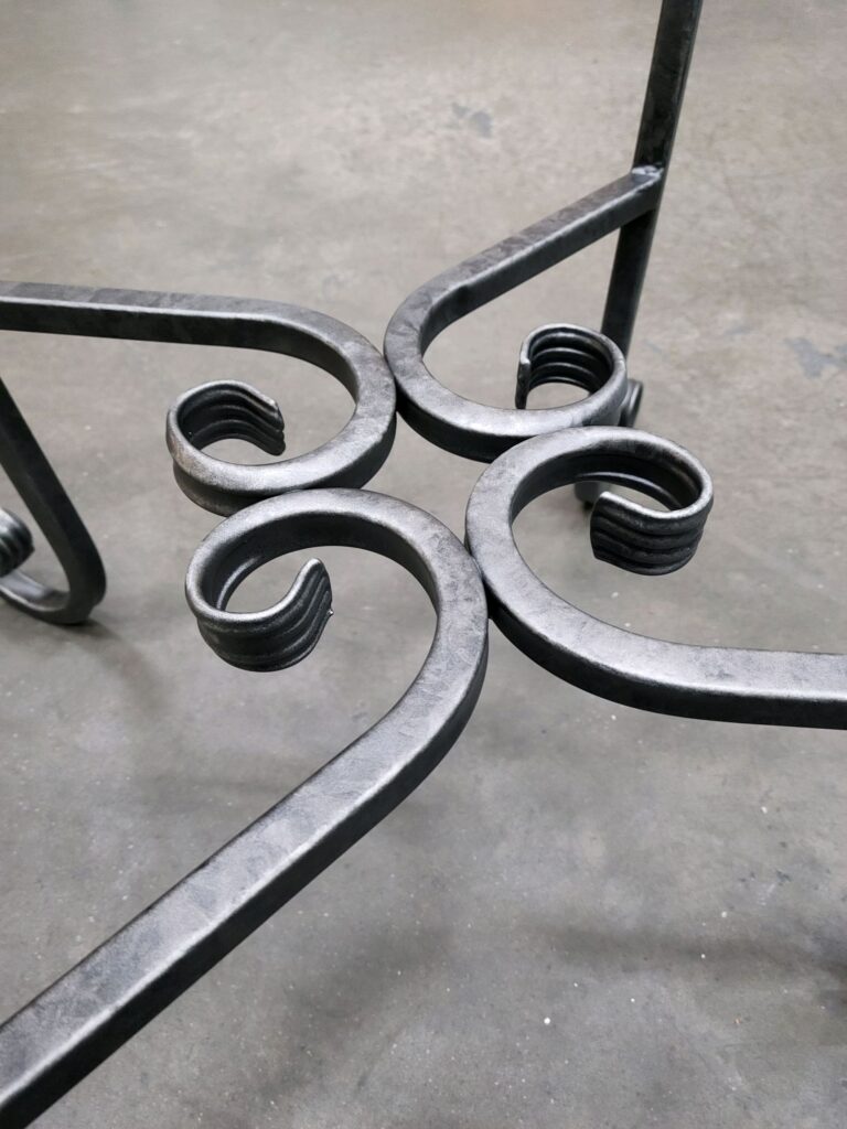 A close-up of a wrought iron table leg with intricate scrollwork, featuring a patina finish that enhances the metal&rsquo;s aged elegance. The smooth yet textured curves display handcrafted artistry in a timeless blackened steel effect.